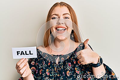 Young beautiful redhead woman holding fall word on paper smiling happy and positive, thumb up doing excellent and approval sign Stock Photo