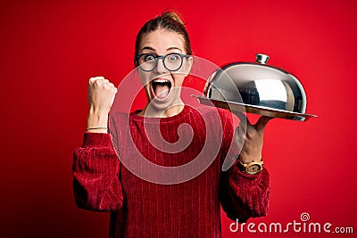 Young beautiful redhead woman holding waitress tray over isolated red background screaming proud and celebrating victory and Stock Photo