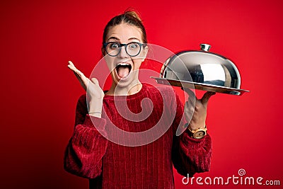 Young beautiful redhead woman holding waitress tray over isolated red background very happy and excited, winner expression Stock Photo