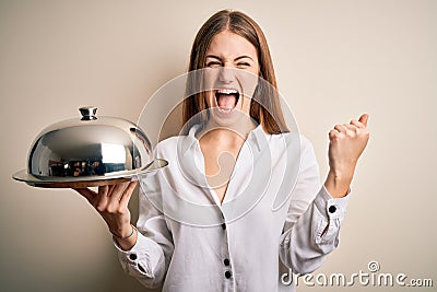 Young beautiful redhead woman holding waitress tray over isolated white background screaming proud and celebrating victory and Stock Photo