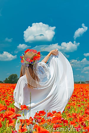 Young beautiful redhead woman in a white shirt with a poppy wreath on her head standing backwards in a poppy field on Stock Photo