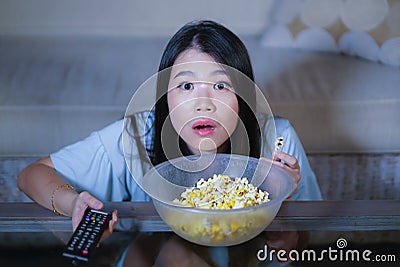 Young beautiful and scared Asian Chinese teenager woman in fear watching horror scary movie at home sofa couch eating popcorn bowl Stock Photo