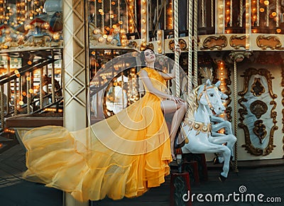 Young beautiful stylish woman sits astride a toy horse, rides a carousel. Long yellow bright dress fluttering in motion Stock Photo