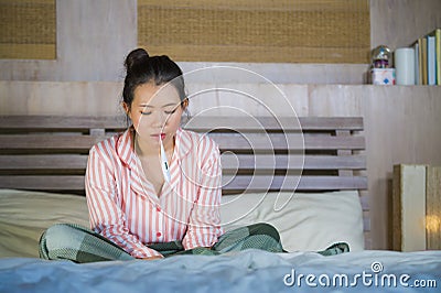 Sweet Asian Korean girl in pajamas covered with blanket sick suffering cold and flu taking temperature with thermometer in bed Stock Photo