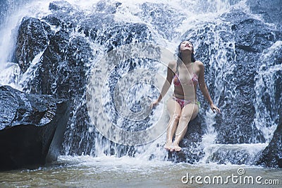 Young beautiful and sweet Asian woman in bikini getting body wet under stream of natural amazing waterfall sitting on rock feeling Stock Photo