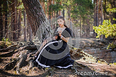 Young, beautiful woman in a black medieval dress with a steel rose in her hands, sitting in the woods on the roots of a tree. Attr Stock Photo