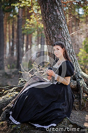 Young, beautiful woman in a black medieval dress with a steel rose in her hands, sitting in the woods on the roots of a tree. Attr Stock Photo