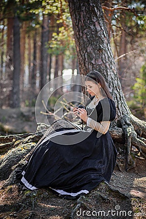 Young, beautiful woman in a black medieval dress with a steel rose in her hands, sitting in the woods on the roots of a tree. Attr Stock Photo