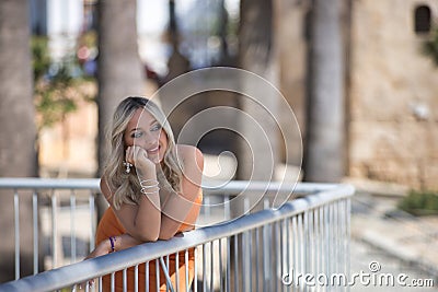 Young and beautiful woman, blonde with blue eyes and an orange dress, leaning on a railing with tender and dreamy look. Concept Stock Photo