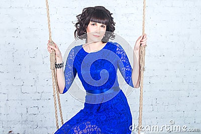 Young beautiful woman in blue dress sitting on swing background of white brick wall. Stock Photo