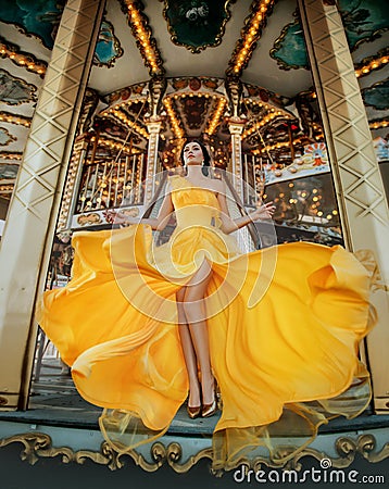 Young beautiful woman in a bright yellow evening dress posing against the backdrop of an amusement park and carousel Stock Photo
