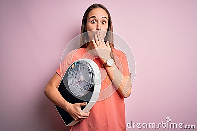 Young beautiful woman controlling weight holding weighting machine over pink background cover mouth with hand shocked with shame Stock Photo