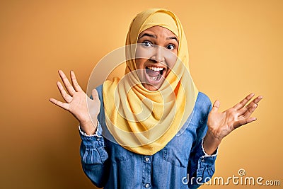 Young beautiful woman with curly hair wearing arab traditional hijab over yellow background celebrating crazy and amazed for Stock Photo