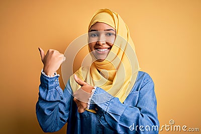 Young beautiful woman with curly hair wearing arab traditional hijab over yellow background Pointing to the back behind with hand Stock Photo