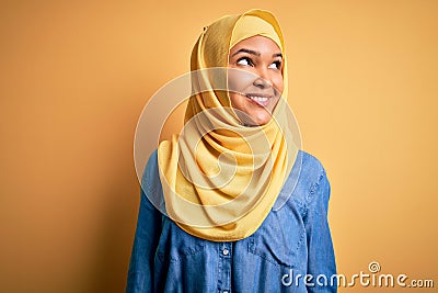 Young beautiful woman with curly hair wearing arab traditional hijab over yellow background smiling looking to the side and Stock Photo