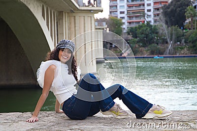 Young, beautiful woman with dark, curly hair and an upturned nose is wearing a cap and sitting by a river. The woman is posing for Stock Photo