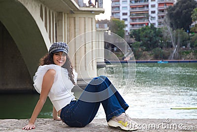 Young, beautiful woman with dark, curly hair and an upturned nose is wearing a cap and sitting by a river. The woman is posing for Stock Photo