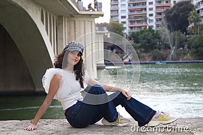 young, beautiful woman with dark, curly hair and an upturned nose is wearing a cap and sitting by a river. The woman is posing for Stock Photo