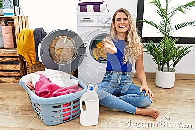 Young beautiful woman doing laundry sitting by wicker basket smiling friendly offering handshake as greeting and welcoming Stock Photo