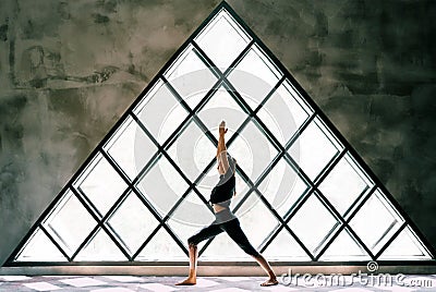 Young beautiful woman doing yoga asana Warrior I Pose on large triangular window background Stock Photo