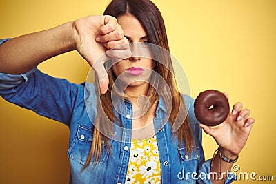 Young beautiful woman eating chocolate donut over yellow background with angry face, negative sign showing dislike with thumbs Stock Photo