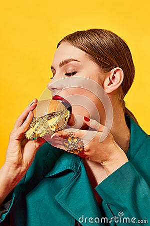 Young beautiful woman in green coat eating, biting cheeseburger with necklaces over yellow background. Luxury food. Food Stock Photo