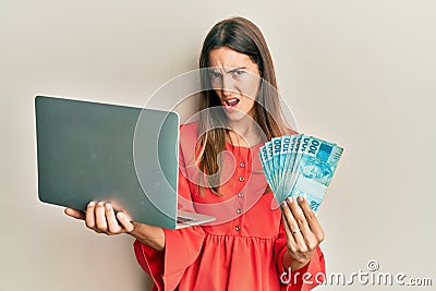 Young beautiful woman holding laptop and holding brazilian real in shock face, looking skeptical and sarcastic, surprised with Stock Photo