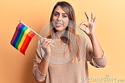 Young beautiful woman holding lgtbq flag doing ok sign with fingers, smiling friendly gesturing excellent symbol Stock Photo