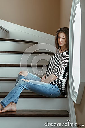 Young beautiful woman model with messy long hair in ripped blue jeans and striped t-shirt sitting indoor by window Stock Photo