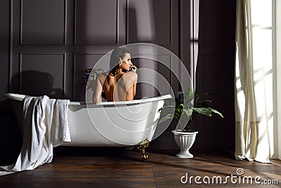 Young beautiful woman sitting in bathroom near expensive bathtub bath looking at the corner on dark Stock Photo