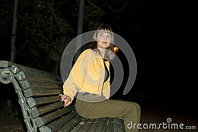 Woman sitting in a bench in a public city park at night while looking to camera Stock Photo