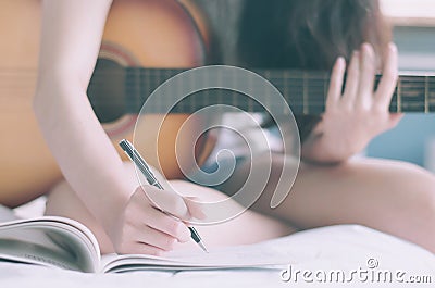 Young beautiful woman sitting on her bed in the bedroom holding guitar composing a song and writing song in textbook ,musician, Stock Photo