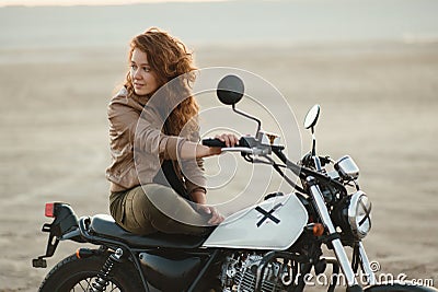 Young beautiful woman sitting on her old cafe racer motorcycle in desert at sunset or sunrise Stock Photo