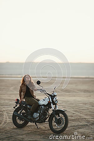 Young beautiful woman sitting on her old cafe racer motorcycle in desert at sunset or sunrise Stock Photo