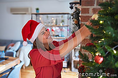Young beautiful woman standing with happy face decoring christmas tree with glass baubles ornaments and wearing santa claus hat Stock Photo