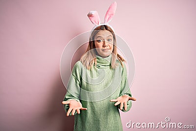 Young beautiful woman wearing easter rabbit ears standing over isolated pink background clueless and confused expression with arms Stock Photo