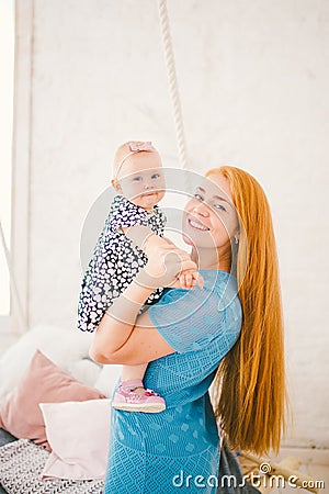 Young beautiful woman with red long hair in a blue dress holds a child on her hands one year blonde near the bed on which lies a m Stock Photo