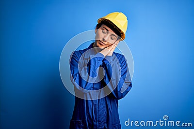 Young beautiful worker woman with blue eyes wearing security helmet and uniform sleeping tired dreaming and posing with hands Stock Photo
