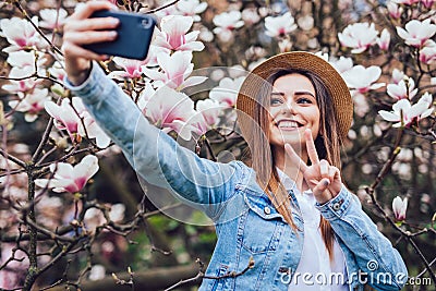 Young beauty woman in summer hat take selfie on phone near blossom magnolia tree in sunny spring day Stock Photo