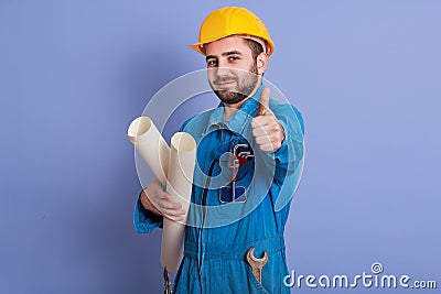 Young beraded caucasian architect with helmet, handsome man posing with thumb up and holding bluepints inhands, wearing blue Stock Photo
