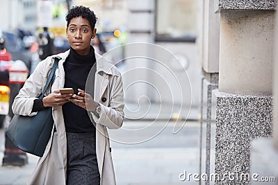 Young black businesswoman walking in the street in London using her smartphone, front view Stock Photo