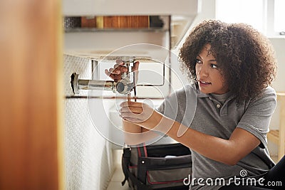 Young black female plumber sitting on the floor fixing a bathroom sink, seen from doorway Stock Photo