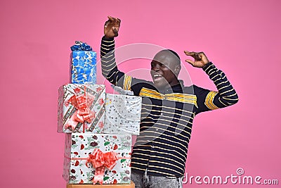 young black man feeling excited and happy and delighted, celebrating while standing next to a stack of gift boxes with fists in Stock Photo