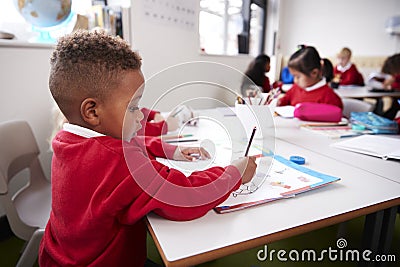 Young black schoolboy wearing school uniform sitting at a desk in an infant school classroom drawing, close up, side view Stock Photo