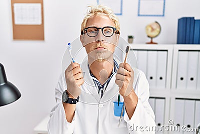 Young blond dentist man working at dentist clinic holding toothbrushes looking at the camera blowing a kiss being lovely and sexy Stock Photo