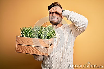 Young blond gardener man with beard and blue eyes holding wooden box with plants stressed with hand on head, shocked with shame Stock Photo