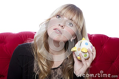 Young blond haired girl on red sofa hears coins in piggybank in Stock Photo