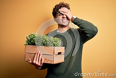 Young blond handsome man with curly hair holding wooden box with plants stressed with hand on head, shocked with shame and Stock Photo