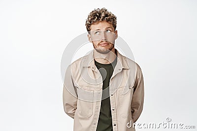 Young blond handsome man looking bored, roll eyes up and exhale, standing with hands behind back against white Stock Photo