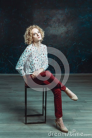 A young blond man androgyne in the image of an attractive woman dressed in a floral shirt, posing sitting on a chair Stock Photo